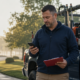 Local Business Owner Pausing Between Jobs | Lucé Media Utility worker in a navy jacket stands by a work truck looking at his smartphone and holding a red clipboard with papers visible near the door